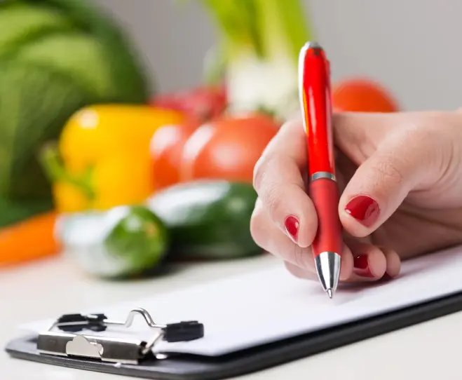 A person writing on paper with vegetables in the background.